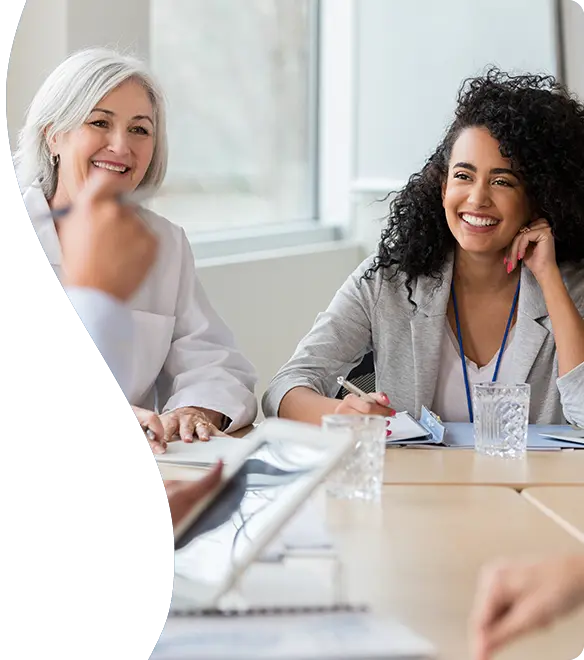 HC20731-senhea-cws-header-584x660px-promanage A group of professionals, including women in business attire and medical uniforms, sit around a conference table engaged in a cheerful and collaborative meeting in a bright, modern office setting.
