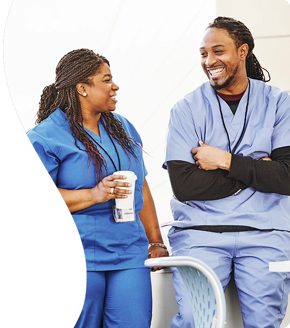 Two healthcare professionals in blue scrubs share a lighthearted conversation and smiles during a break, with one holding a coffee cup in a casual, relaxed setting.