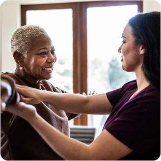 A woman smiling as a healthcare worker helps her with arm exercises in a bright, sunlit room.