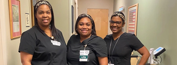 Three smiling healthcare workers in matching black scrubs and headbands stand together in a hallway.