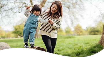 A woman helping a female toddler balance on a fallen tree outdoor on a sunny day.