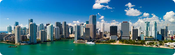 Panoramic view of Miami’s downtown skyline with tall buildings along the turquoise waterfront under a bright blue sky.