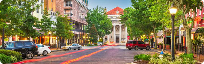 Charming downtown street in North Central Florida with trees, historic buildings, and light trails from passing cars at dusk.