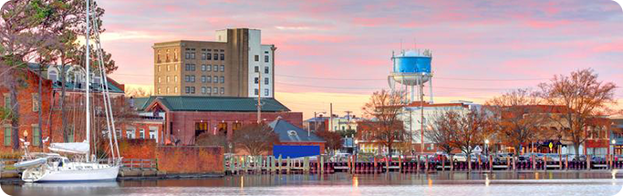 Waterfront view of a North Carolina town at sunset, with boats, buildings, and a blue water tower under a pastel sky.