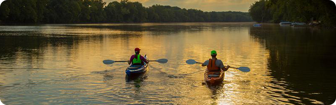 Two people kayak on a calm river at sunset, surrounded by trees and golden reflections on the water.