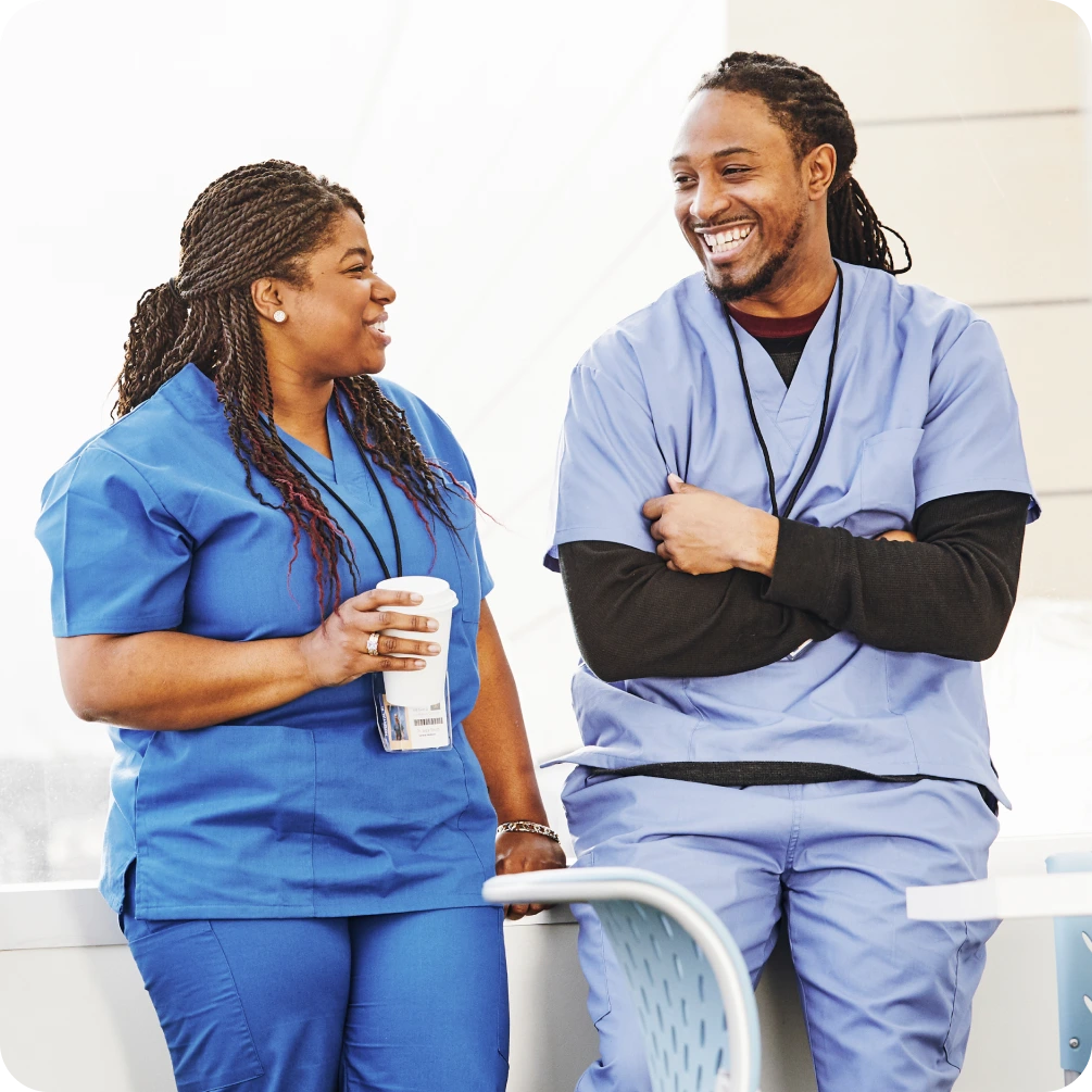Two nurses in scrubs smiling and chatting during a break, one holding a coffee cup in a bright hospital setting.