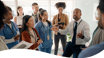 STUD360-senhea-cws-mobile-header-360x200px-supportservices (1) A man in business attire speaks to a group of healthcare professionals, some in scrubs and others in business clothing, gathered closely in a bright office setting.