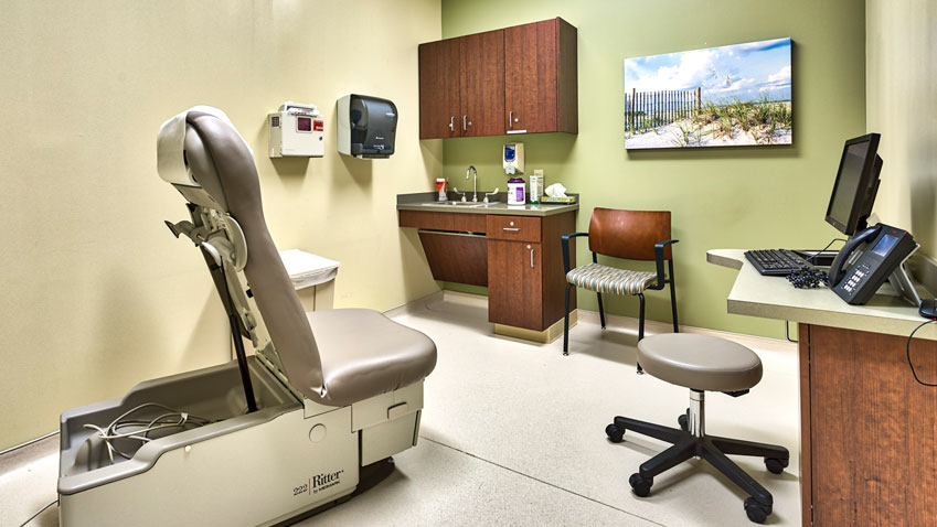 A clean and organized medical exam room with an adjustable chair, desk, computer, and medical supplies.